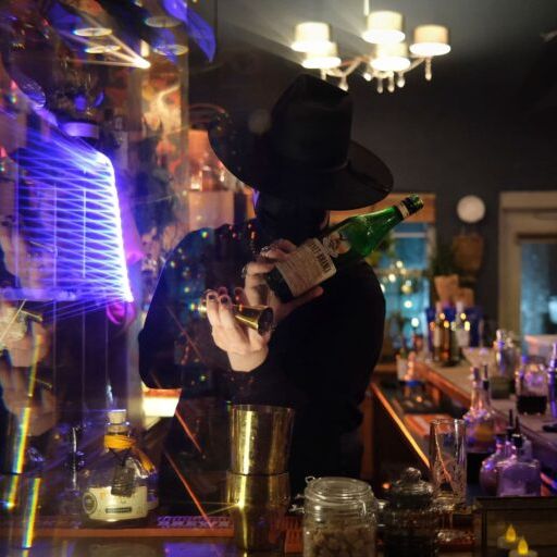 Photo of bartender wearing a hat pouring drink into a cup. In the background, glasses and bottles.
